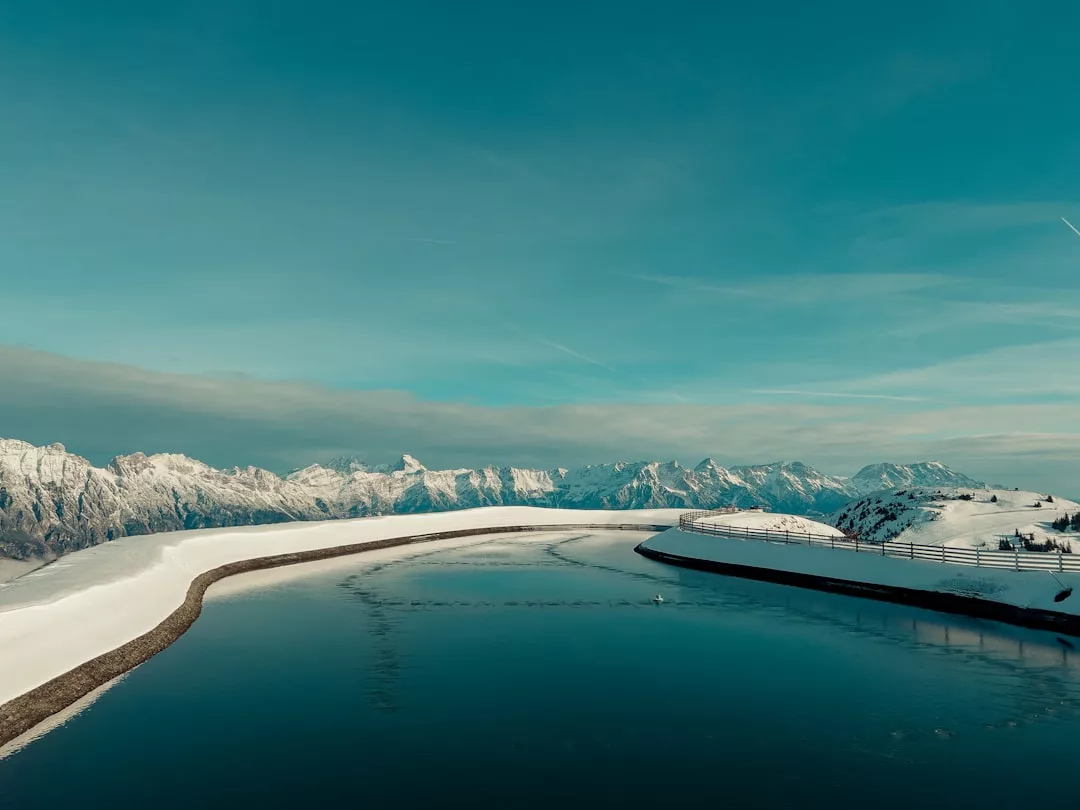 Un embalse tranquilo y curvado bordeado de nieve evoca el lujo de Lujo, mientras que las lejanas montañas nevadas bajo un cielo azul despejado insinúan la tranquila belleza de los serenos paisajes invernales de Mallorca.