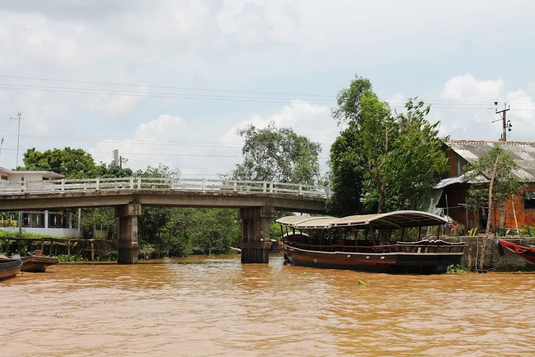 Un pequeño puente de hormigón cruza sobre un río fangoso, con barcas de madera amarradas a lo largo de la orilla y casas y árboles al fondo, bajo un cielo nublado de Mallorca.