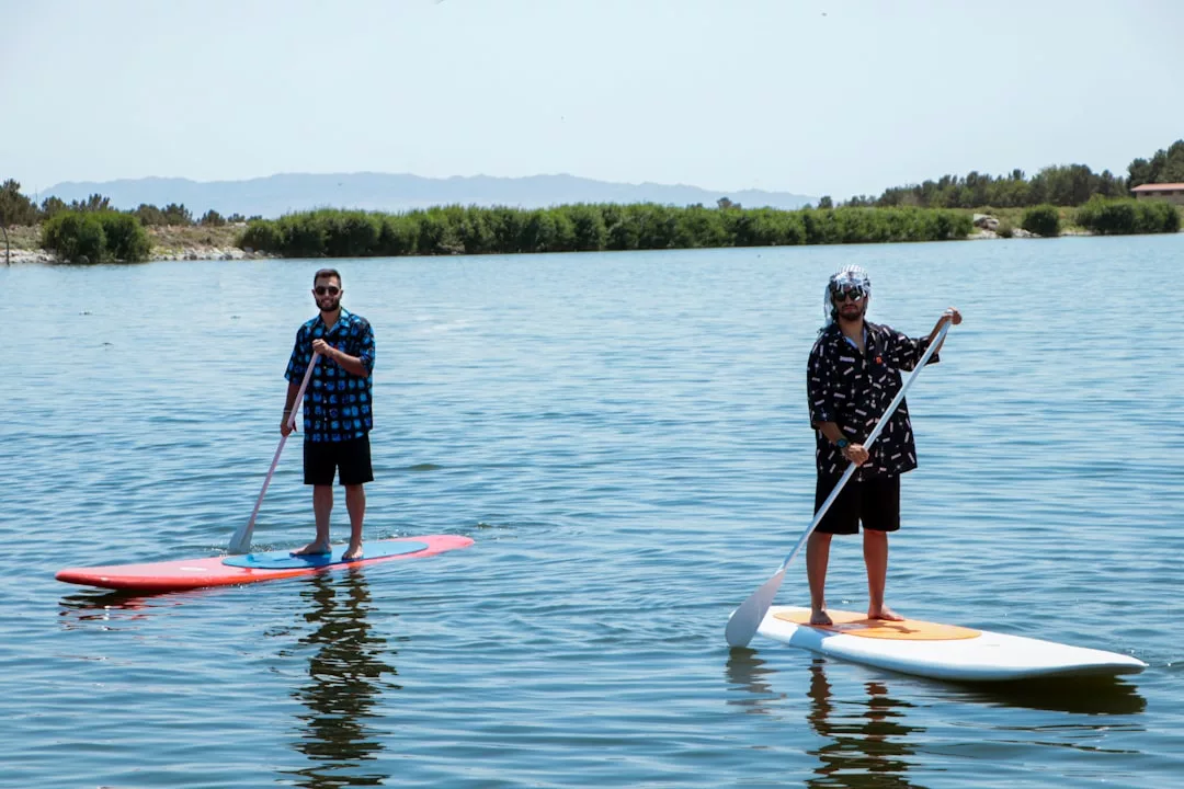 Dos personas subidas a una tabla de paddle surf en un tranquilo lago de Mallorca, con camisetas estampadas y pantalones cortos. Con árboles y colinas a sus espaldas, disfrutan de un día de lujo rodeados de la belleza de la naturaleza.