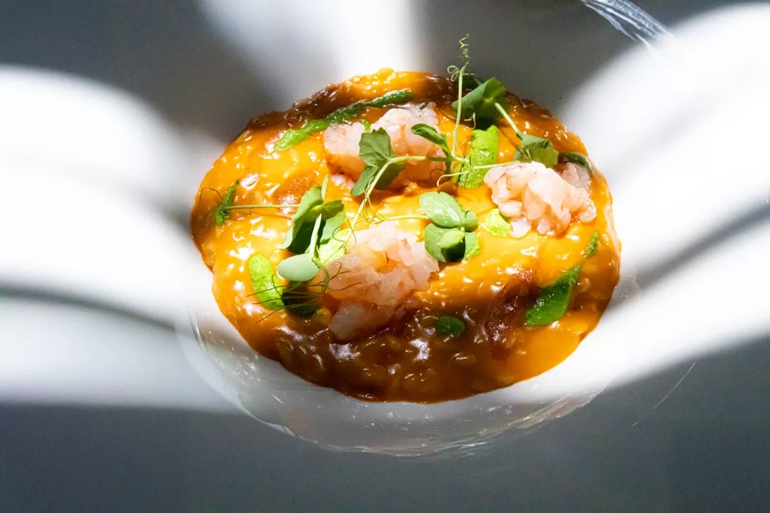 Un cuenco de risotto anaranjado adornado con hierbas verdes y trozos de gamba cruda, iluminado por dramáticas luces y sombras, evoca el lujo de la vibrante escena culinaria de Mallorca.