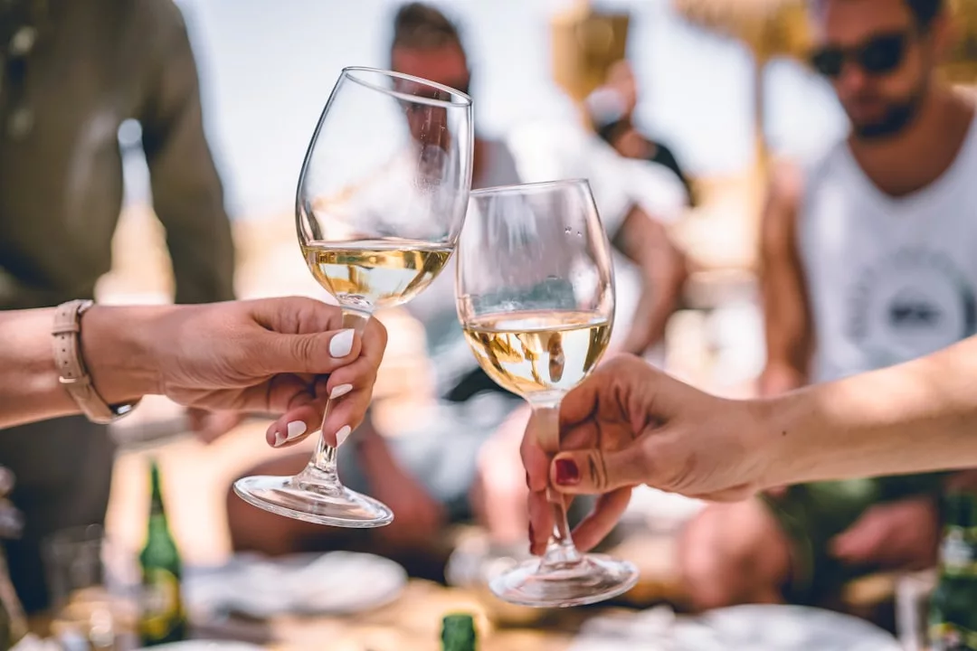 Dos personas brindan con vino blanco al aire libre en Mallorca, mientras otras se relajan en el fondo borroso en un día soleado, captando un momento de puro lujo.