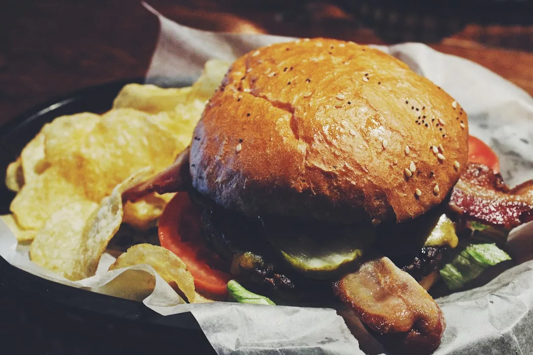 Una hamburguesa de bacon y queso con lechuga, tomate, pepinillos y un panecillo de sésamo, servida con una guarnición de patatas fritas en papel de pergamino, perfecta para saborear el lujo tanto si estás cenando en Mallorca como en casa.