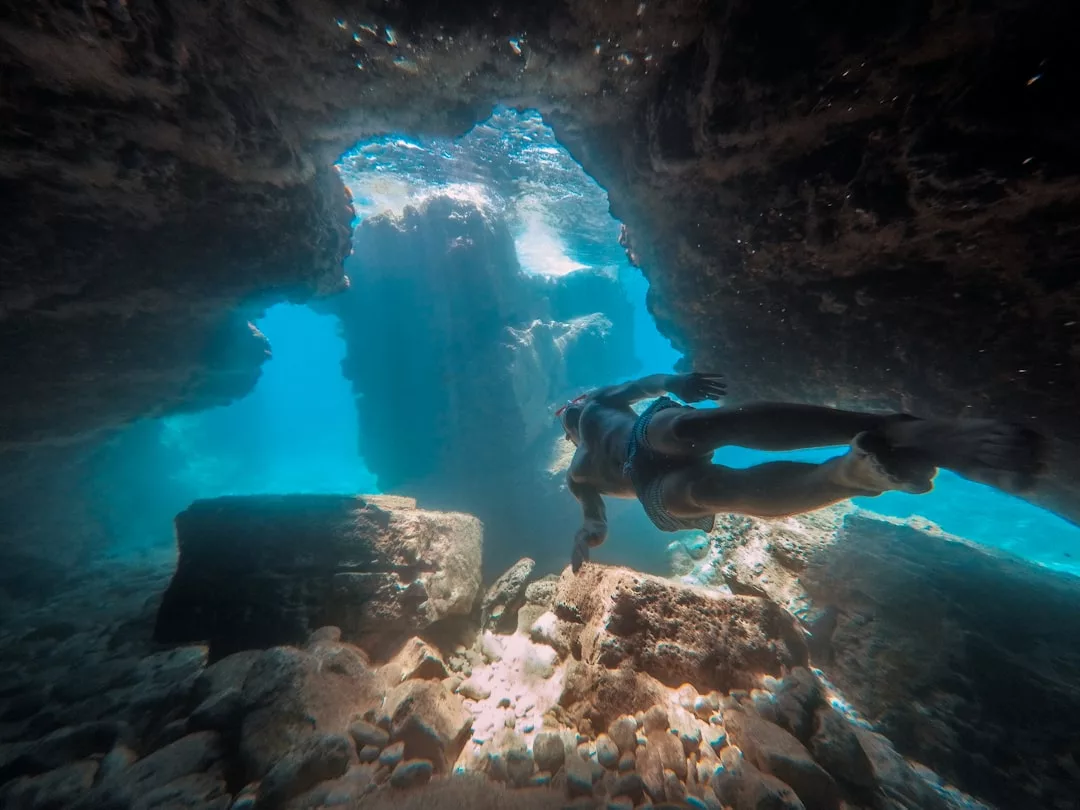 Una persona en bañador está bajo el agua, nadando a través de una formación rocosa en una cueva de Mallorca, con la luz del sol entrando desde arriba.