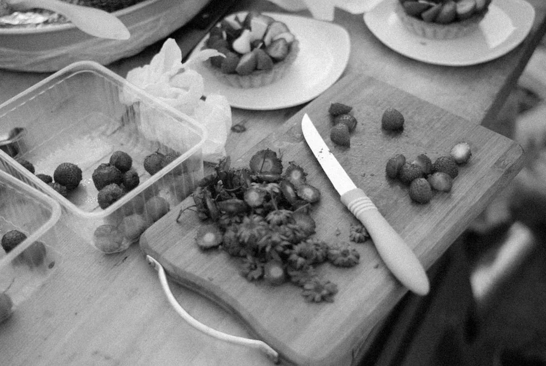Una tabla de cortar con un cuchillo, tapas de fresas y fresas en rodajas se encuentra junto a recipientes de plástico y platos con tartas sobre una superficie de madera, evocando el lujo de una pastelería de inspiración mallorquina.