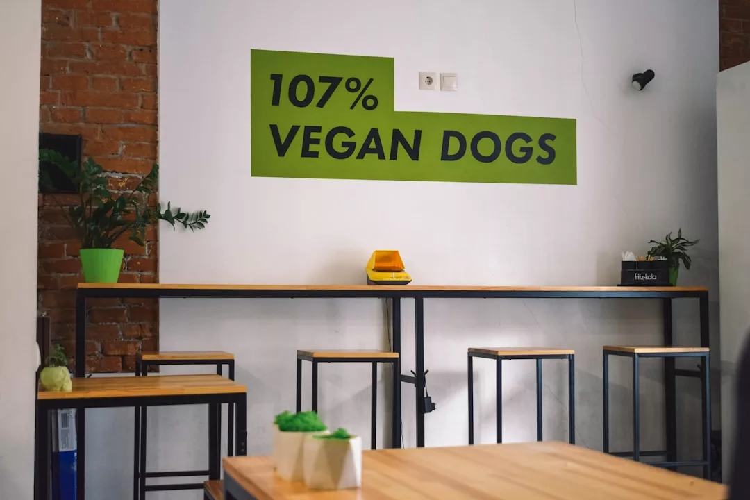 El interior de una cafetería minimalista de Mallorca, con mesas y taburetes de madera, exuberantes plantas verdes en macetas y un letrero en la pared que reza "107% VEGAN DOGS", ofrece un toque de lujo en cada detalle.