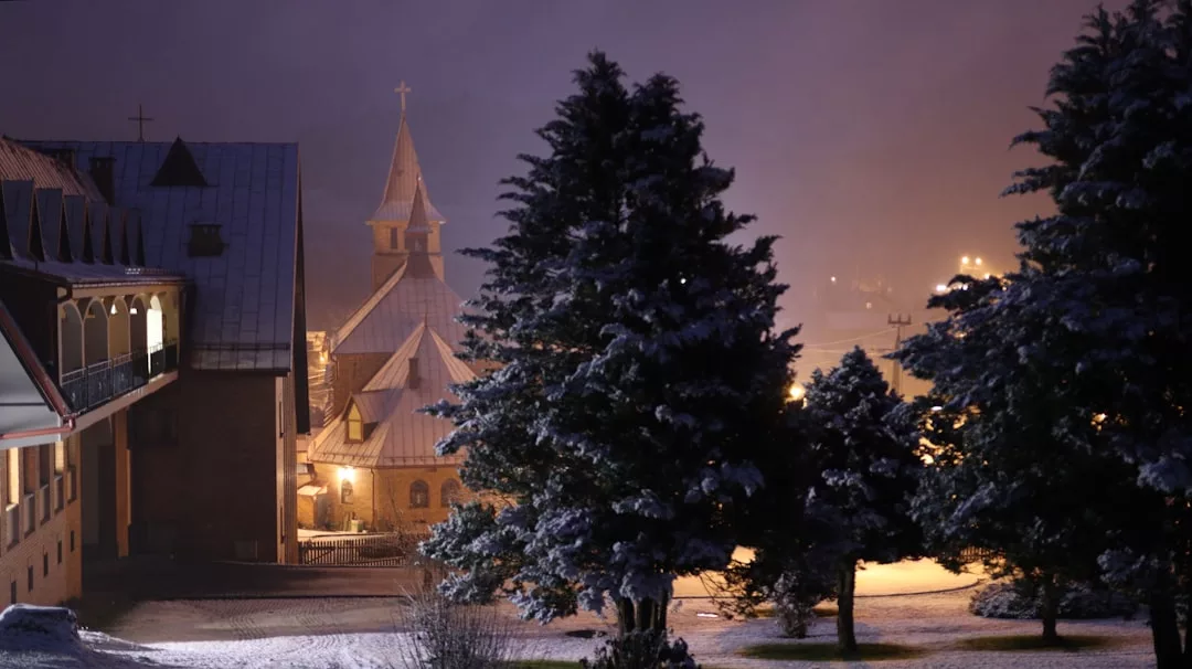 La escena nocturna de un pueblo nevado con edificios iluminados y una iglesia, rodeada de altos pinos espolvoreados de nieve, evoca una sensación de lujo que recuerda a los encantadores retiros invernales de Mallorca.