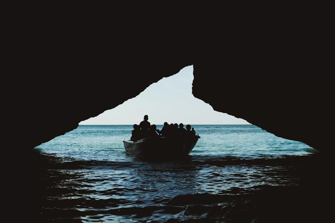 La silueta de una barca que transporta personas sale de una oscura cueva marina de Mallorca, en dirección a aguas abiertas bajo un cielo brillante.