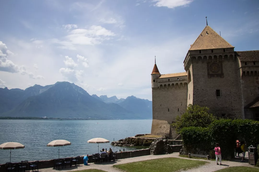 Un castillo de piedra se alza junto a un lago con montañas al fondo; la gente pasea cerca y se sienta bajo las sombrillas a lo largo de la orilla, disfrutando de un aire de lujo que recuerda a los tranquilos retiros de Mallorca.