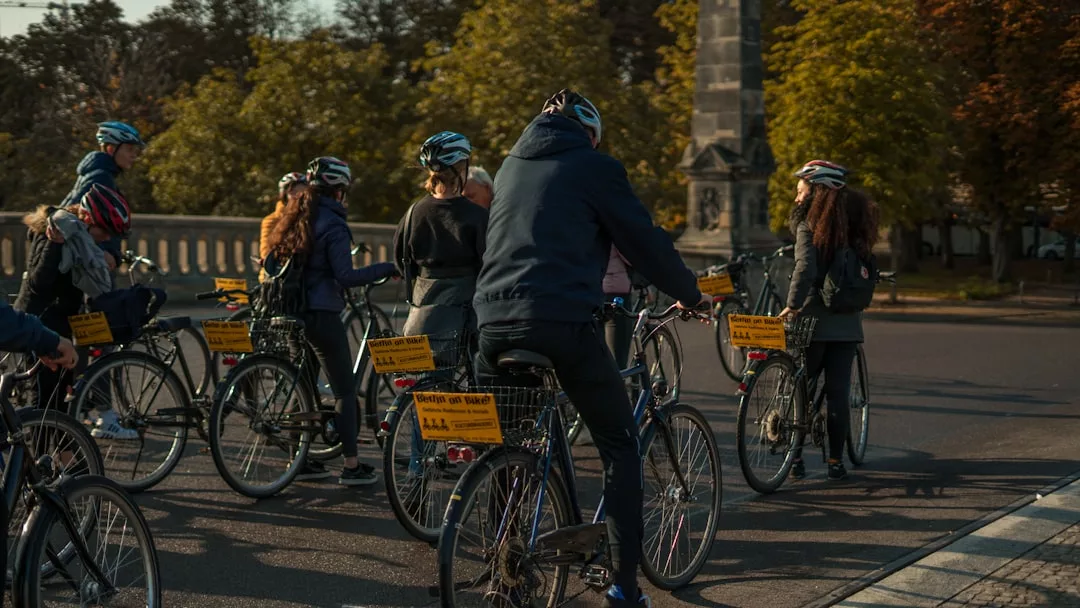Un grupo de personas con casco montan juntas en bicicleta por una calle asfaltada de Mallorca, con árboles y un monumento de piedra que añaden un toque de lujo al pintoresco fondo.