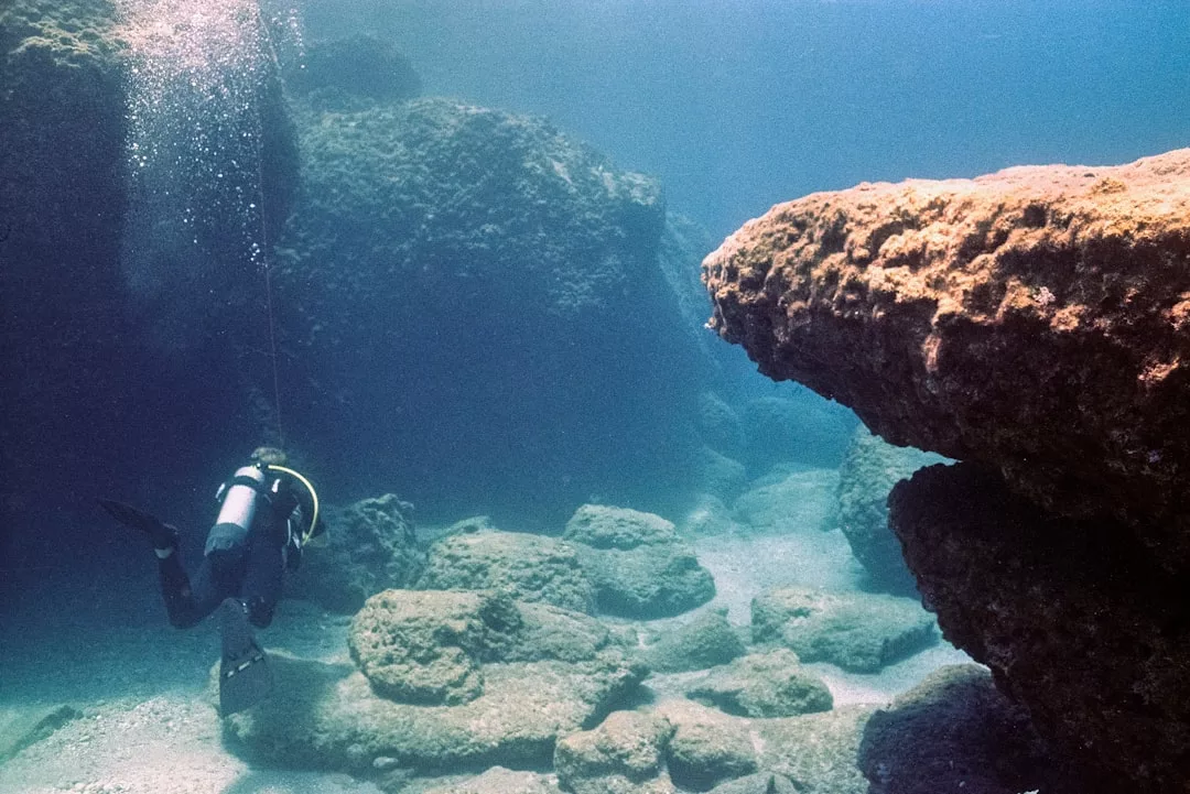 Un submarinista nada cerca de grandes rocas y peñascos submarinos frente a la costa de Mallorca, con la luz del sol atravesando el agua, creando una escena de lujo natural.
