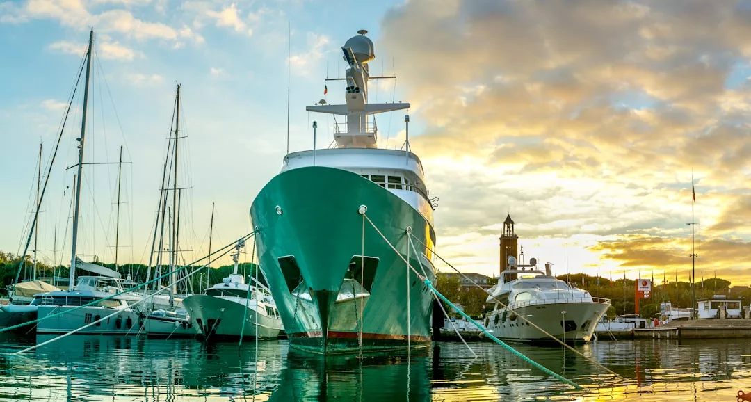 Gran yate de casco verde atracado en un puerto deportivo de Mallorca, rodeado de otras embarcaciones, con un cielo al atardecer y una torre del reloj al fondo, captando la esencia del lujo en la isla.