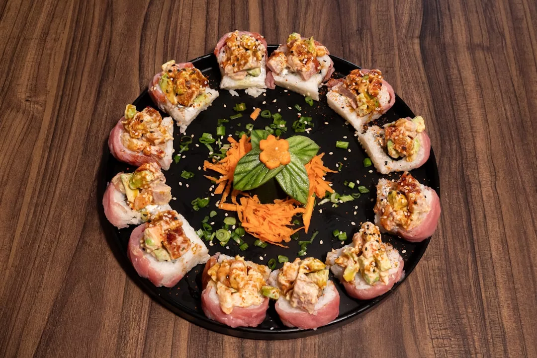 Un plato negro con un anillo de rollos de sushi cubiertos con una cremosa mezcla de marisco, adornado con cebollas verdes, zanahorias ralladas y una flor de pepino en el centro: un arreglo que aporta un toque de Lujo a su experiencia gastronómica inspirada en Mallorca.