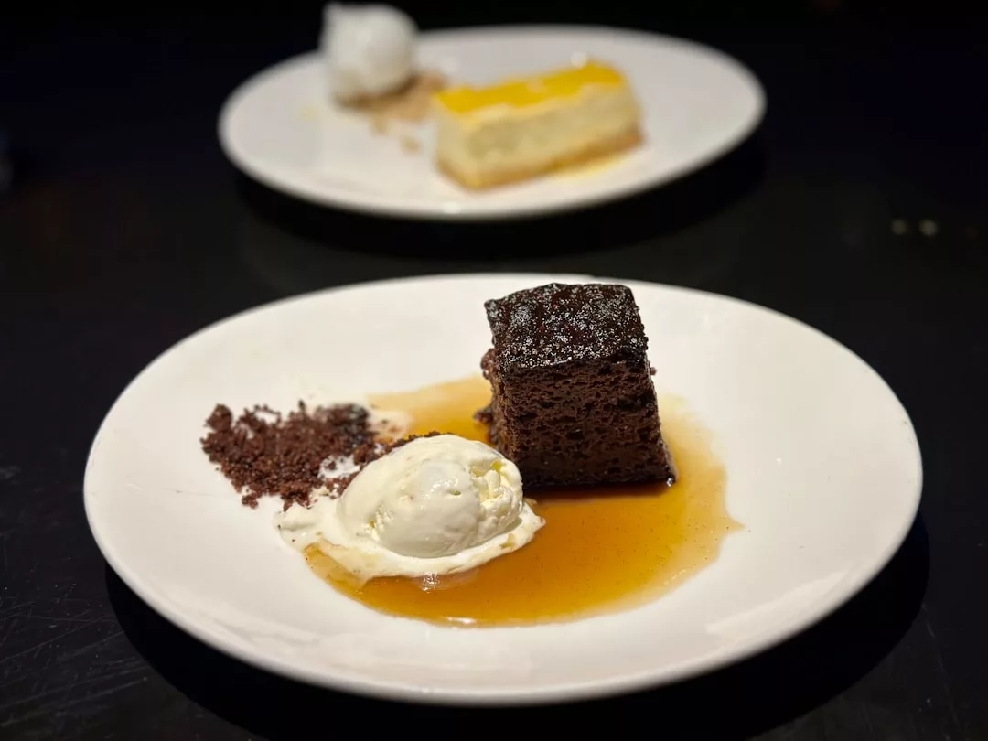 Un plato de postre con un trozo de tarta de chocolate, una bola de helado de vainilla, migas de chocolate y salsa encarna el lujo de Mallorca; otro postre aparece difuminado al fondo.