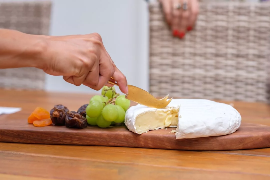 Una mano usa un cuchillo para cortar queso blanco suave sobre una tabla de madera, acompañado de uvas verdes, albaricoques secos y dátiles: un toque de lujo mallorquín en cada bocado.