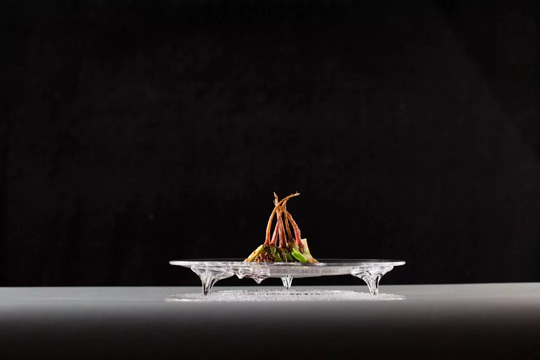 Un plato gourmet pequeño y artísticamente emplatado se centra en un plato de cristal transparente y elevado sobre un fondo negro, una presentación característica que recuerda a la escena gastronómica de Lujo en Mallorca.