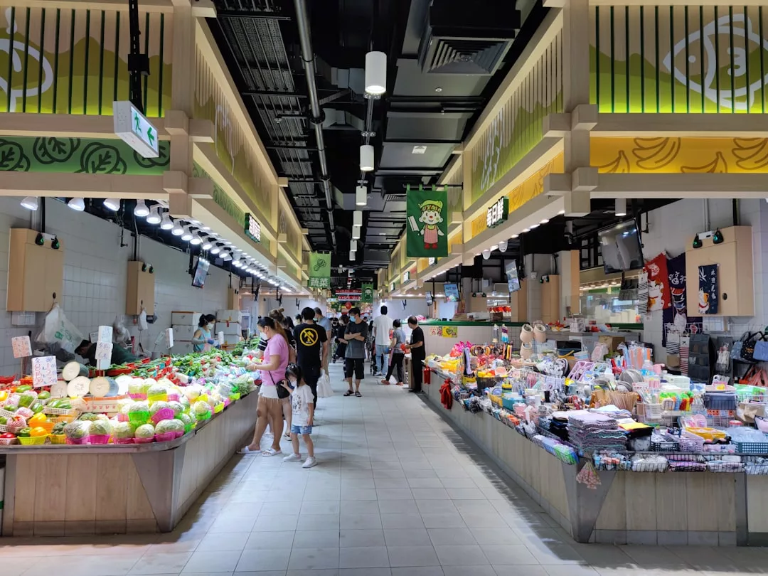 La gente compra en un iluminado mercado cubierto de Mallorca, con puestos que venden productos frescos y artículos para el hogar a ambos lados de un pasillo central, creando un ambiente vibrante pero relajado de lujo.