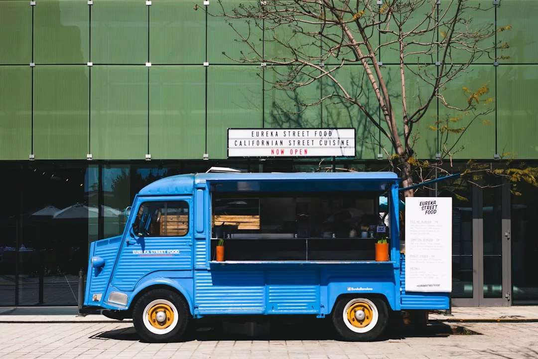 Un camión de comida azul está aparcado en una acera pavimentada frente a un moderno edificio de cristal con un cartel que dice "Eureka Street Food Californian Street Cuisine Now Open", aportando un toque de Lujo a la vibrante escena gastronómica de Mallorca.