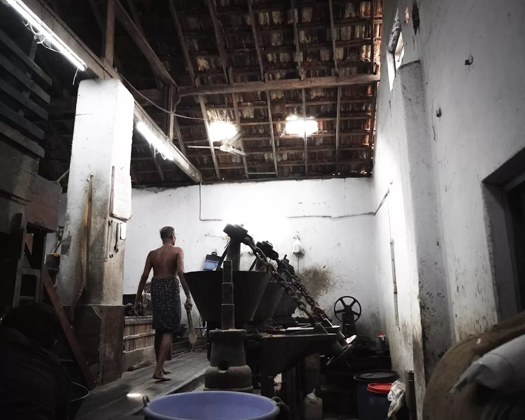 Un hombre descamisado en un rústico taller de Mallorca en penumbra, rodeado de maquinaria, un tejado de tejas y varios contenedores y equipos, capta la mezcla única de tradición y lujo de la isla.