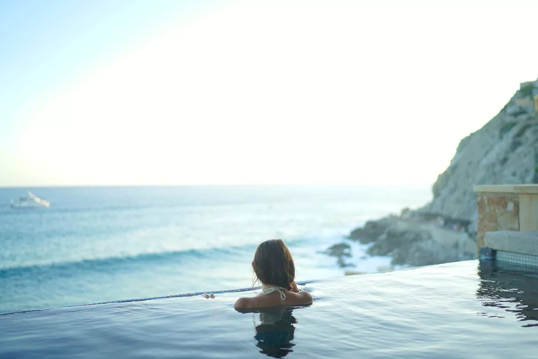 Una persona se relaja en una piscina infinita con vistas al océano en Mallorca, con olas y acantilados rocosos visibles al fondo bajo un cielo luminoso, disfrutando del verdadero lujo junto al mar.