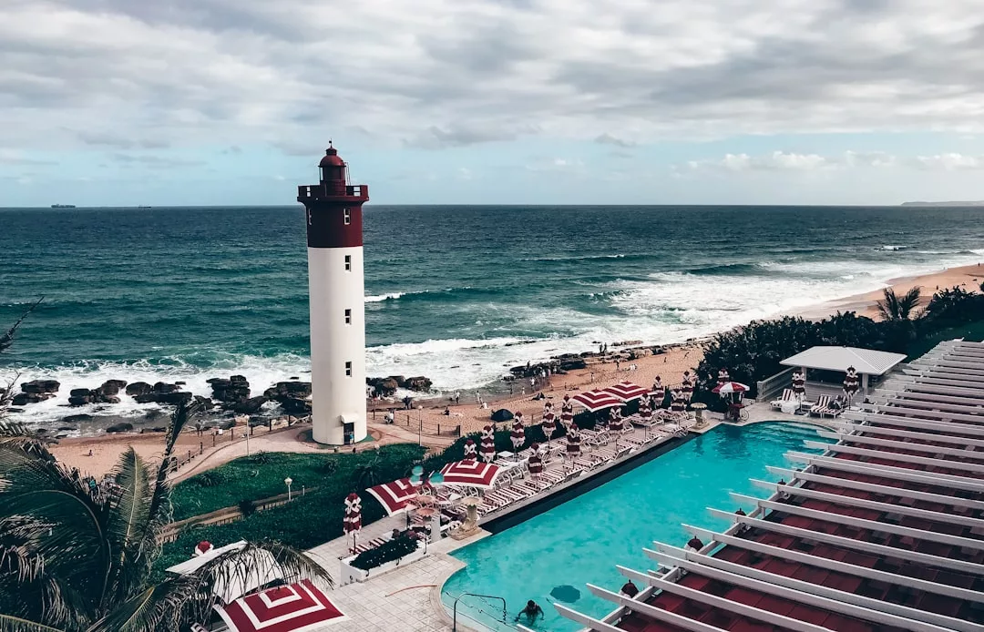 Un faro se alza cerca del océano en una playa de arena de Mallorca, con una piscina, tumbonas y sombrillas de rayas rojas en primer plano, ofreciendo un verdadero toque de lujo bajo un cielo nublado.