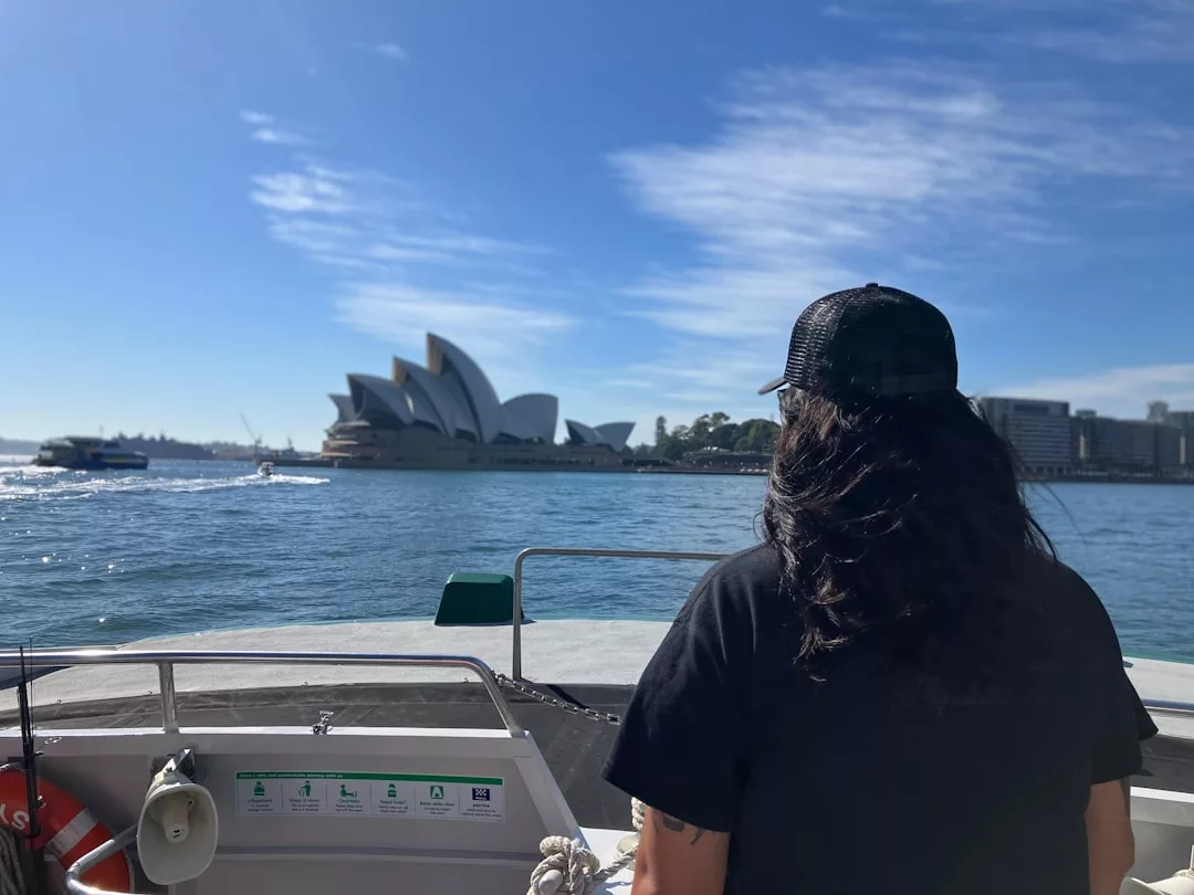 Una persona con el pelo largo y oscuro y una gorra negra está de pie en un barco, disfrutando de un soleado día de lujo mientras mira hacia la Ópera de Sydney al otro lado del agua, que recuerda a las hermosas costas de Mallorca.
