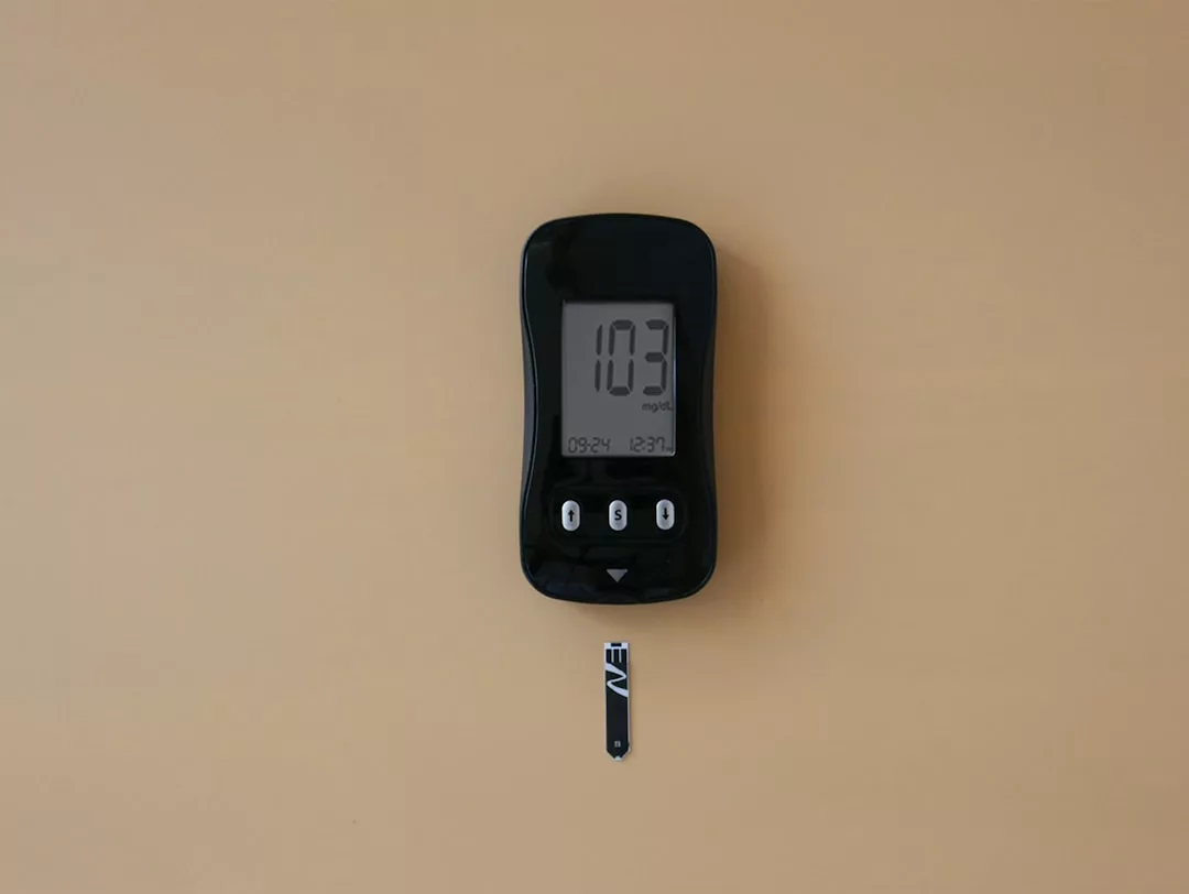 Un medidor de glucosa negro que muestra una lectura de 103 mg/dL, con una tira reactiva insertada, colocado sobre un fondo beige, evoca el discreto lujo del bienestar diario, como las serenas mañanas de Mallorca.