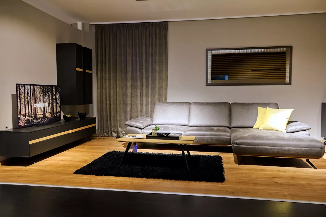 Moderno salón con un sofá seccional gris, cojín amarillo, mesa de centro de madera, televisor en una consola, alfombra negra y una gran cortina, que ofrece un toque de lujo inspirado en Mallorca para un confort con estilo.