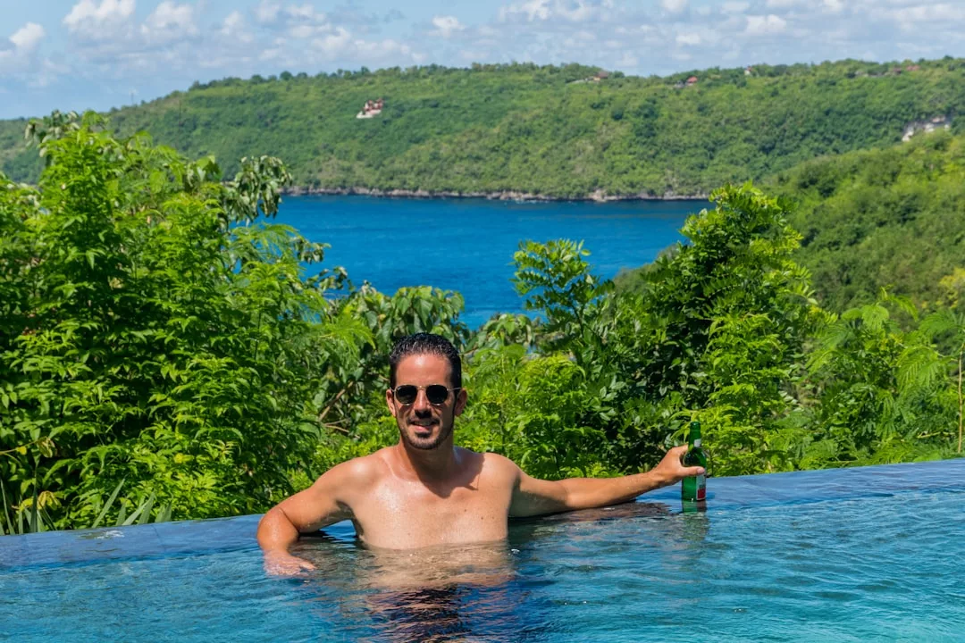 Un hombre con gafas de sol se sienta en una piscina infinita con una botella en la mano, disfrutando del lujo y de las impresionantes vistas de exuberantes colinas verdes y aguas azules de fondo, que recuerdan al impresionante paisaje de Mallorca.