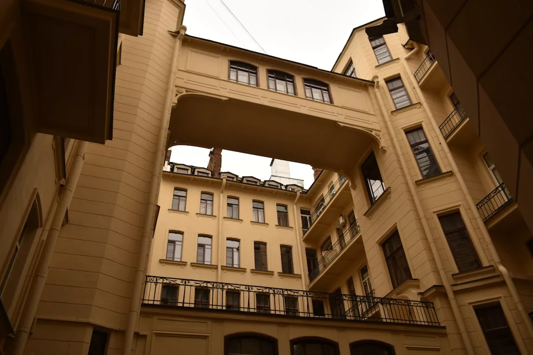 Vista de un edificio beige de varias plantas con un puente elevado cerrado que conecta dos secciones, visto desde un patio inferior, que destila el lujo refinado y la elegancia que a menudo se encuentran en las joyas arquitectónicas de Mallorca.