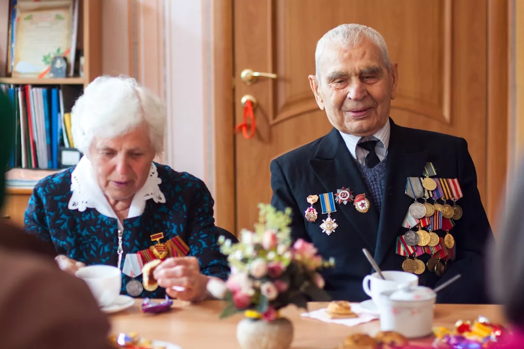 Un hombre y una mujer mayores con medallas prendidas en sus chaquetas se sientan a una mesa con té y aperitivos, disfrutando de un toque de lujo, con un jarrón de flores en primer plano, que recuerda a una tranquila tarde en Mallorca.