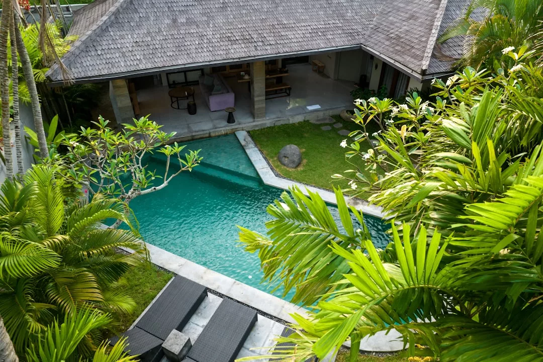 Vista de una piscina turquesa rodeada de plantas tropicales junto a una villa con tejado de paja y patio abierto, que capta la esencia del lujo y la relajación en la hermosa Mallorca.