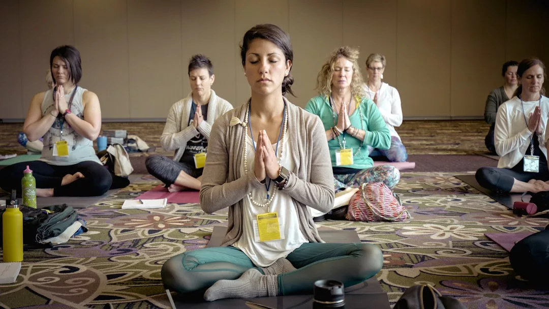 Un grupo de personas sentadas con las piernas cruzadas sobre esterillas de yoga en un interior de Mallorca, con los ojos cerrados y las manos en posición de oración, disfrutan de una sesión de meditación de lujo.