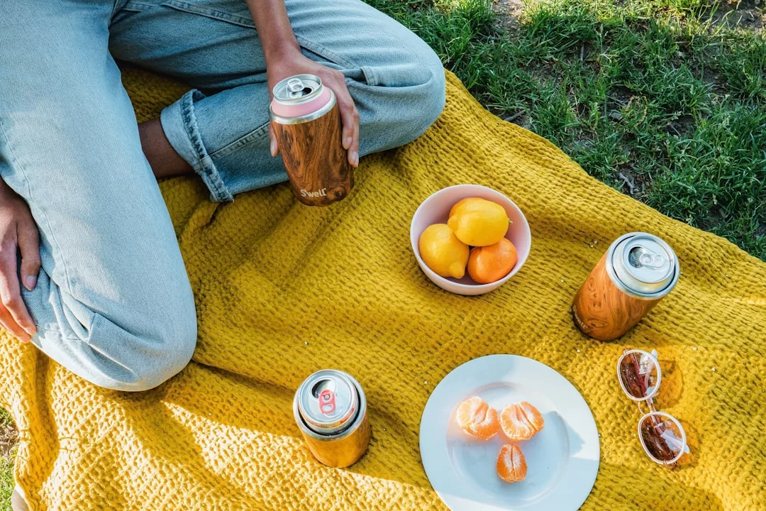 Disfrutando de un toque de lujo en Mallorca, una persona se relaja en una manta amarilla al aire libre con latas, limones y naranjas frescos, rodajas de naranja peladas y gafas de sol esparcidas por la hierba.