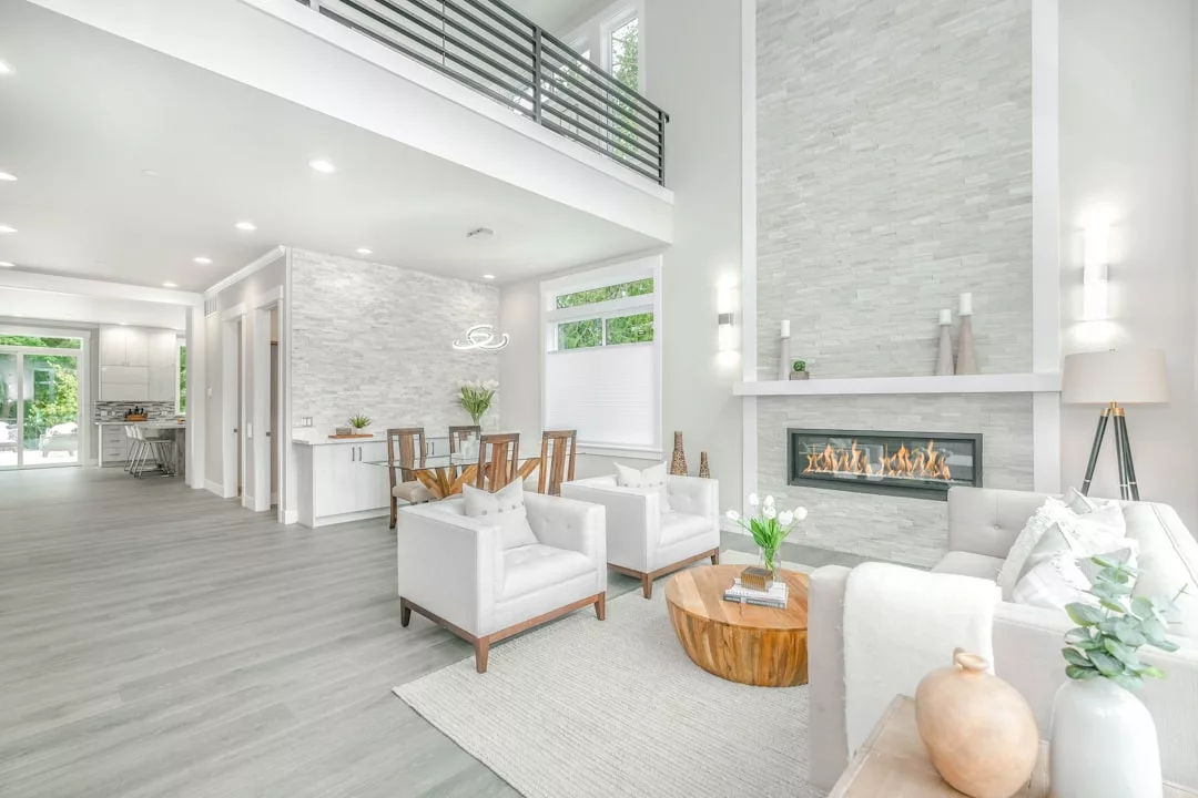 Moderno salón de concepto abierto con muebles de color gris claro, chimenea de piedra, detalles de madera y grandes ventanales que llenan el espacio de luz natural. Disfrute de un toque de lujo inspirado en Mallorca en la zona de comedor visible y la cocina al fondo.