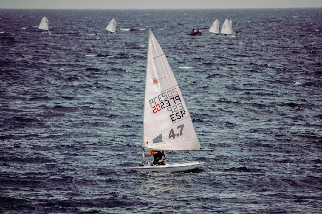 Una persona navega en un pequeño bote blanco con una vela numerada en aguas abiertas de Mallorca, con varios otros veleros al fondo, captando el espíritu de lujo y aventura.