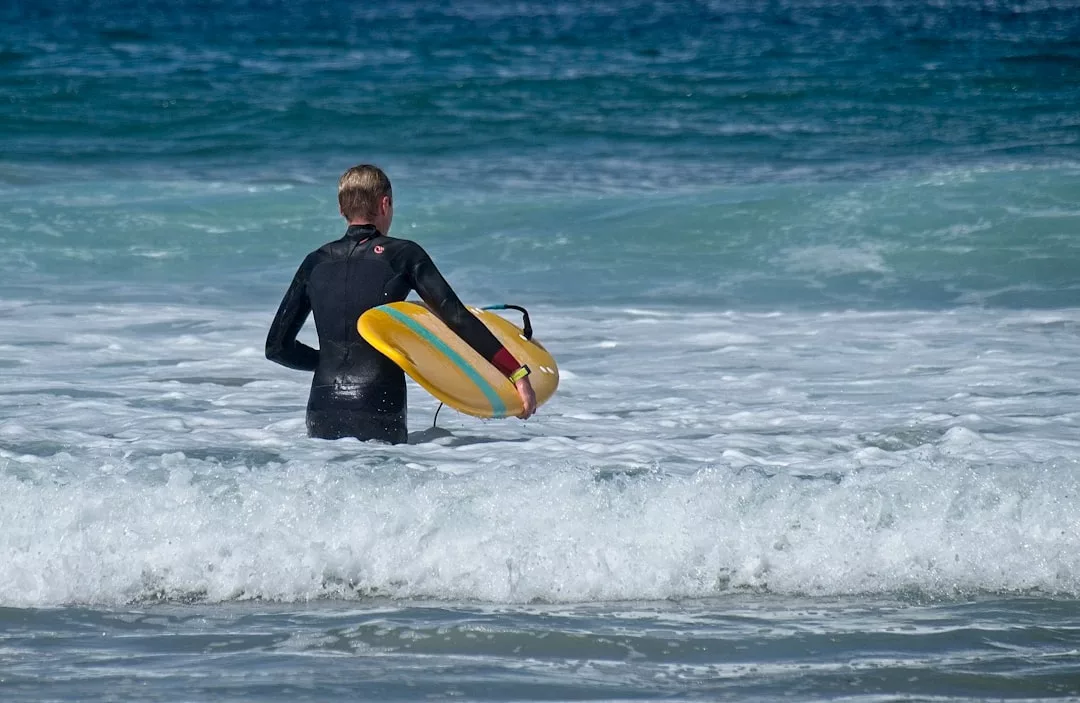 Una persona con traje de neopreno se encuentra en aguas poco profundas del océano sujetando una tabla de surf amarilla, mirando hacia las olas, dispuesta a disfrutar del lujo del surf en la pintoresca costa de Mallorca.