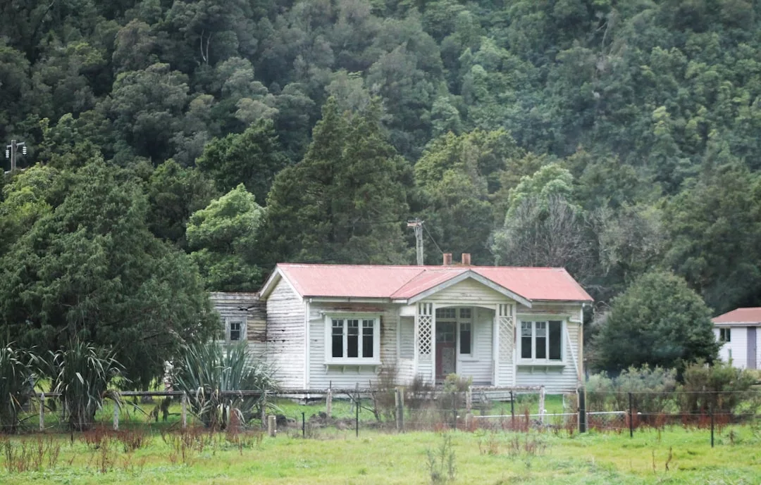 Una pequeña casa blanca y desgastada con tejado rojo, rodeada de árboles y vegetación en Mallorca, destila encanto rústico y un discreto lujo, con una valla en primer plano.