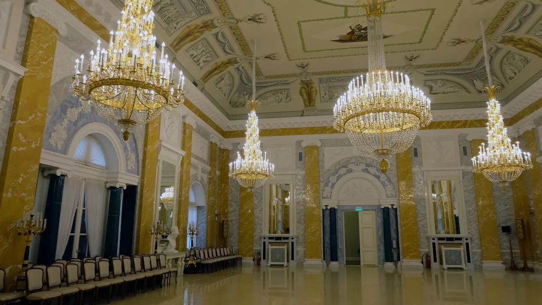 Un gran salón de baile con paredes de mármol, lámparas de araña ornamentadas, columnas decorativas e hileras de sillas tapizadas evoca el lujo intemporal de Mallorca a través de sus altas ventanas arqueadas y su elegante ambiente.