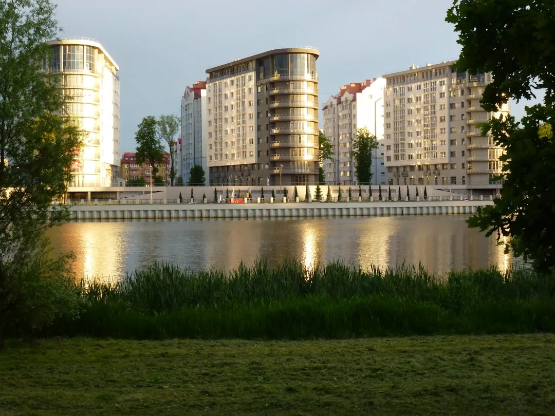 Varios modernos edificios de apartamentos de lujo se reflejan en un río tranquilo, con hierba verde y árboles en primer plano, evocando la serena belleza de la vida mallorquina.