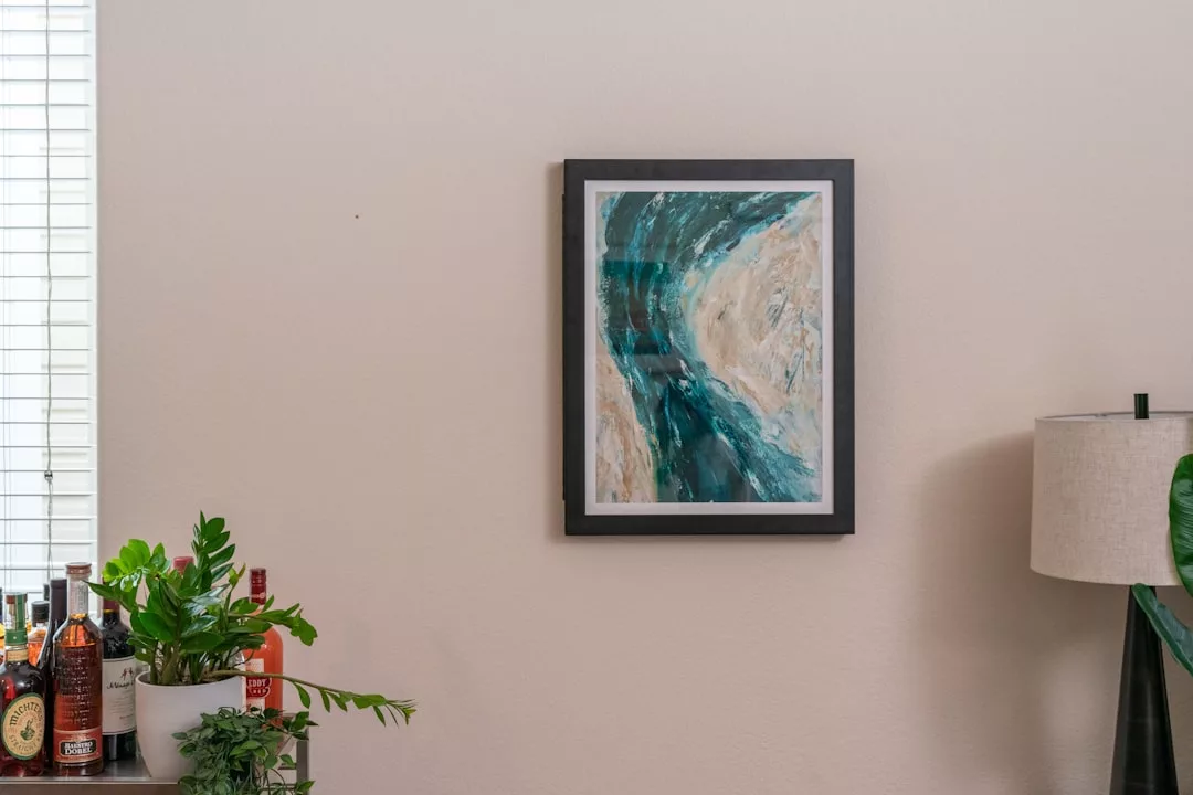 Un cuadro abstracto en tonos azules y beige enmarcado en una pared clara evoca el encanto costero de Mallorca, colocado junto a un alféizar con plantas, botellas y un toque de lujo de la elegante lámpara de pie de la derecha.
