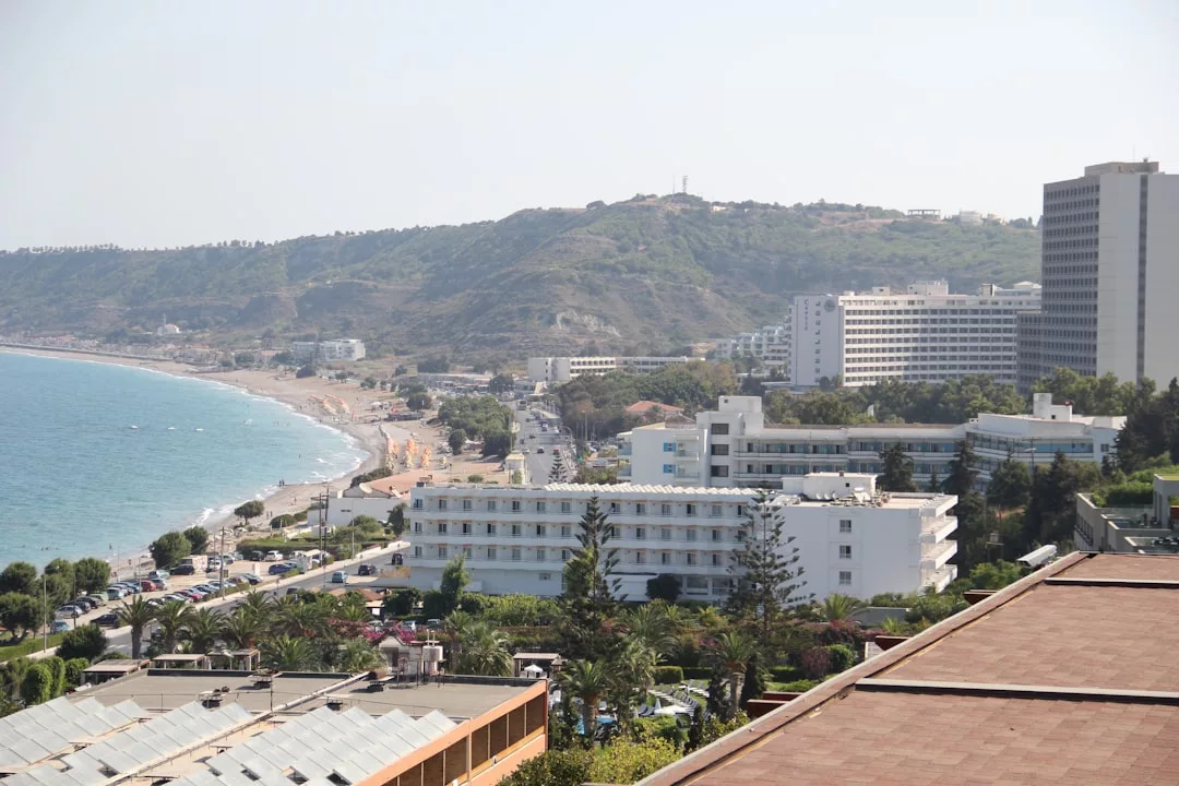 Un paisaje urbano costero en Mallorca con varios hoteles y edificios de mediana altura a lo largo de una playa, bordeada de verdes colinas y el mar bajo un cielo brumoso, desprende un aire de Lujo.