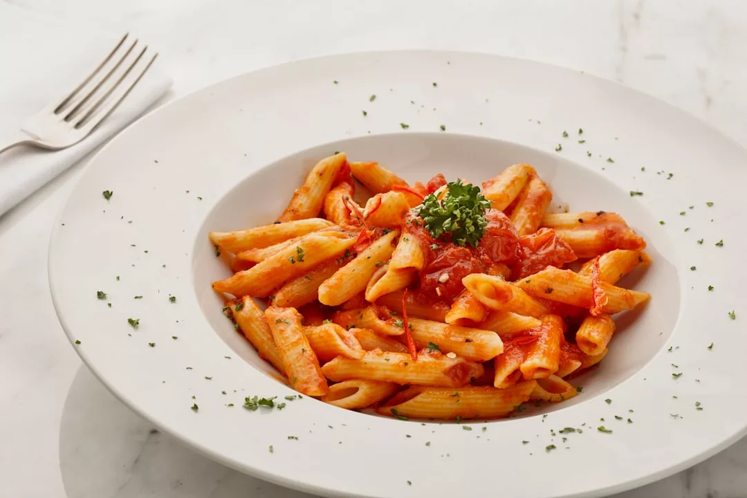 Un plato de pasta penne con salsa de tomate, adornado con perejil picado y una ramita de perejil, ofrece un toque de Lujo mientras reposa en una mesa blanca de Mallorca, junto a una servilleta doblada y un tenedor.
