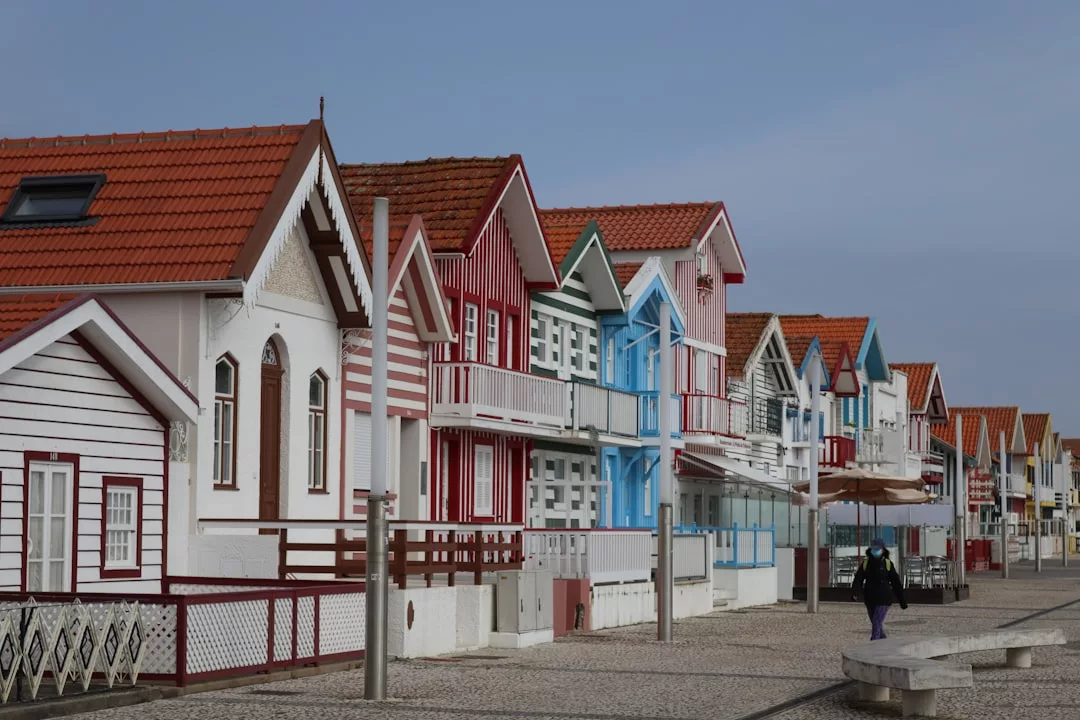 Una hilera de coloridas casas de playa a rayas con tejados de tejas rojas se alinean en un paseo pavimentado bajo el cielo despejado de Mallorca; una persona camina en primer plano, captando un toque de lujo junto al mar.