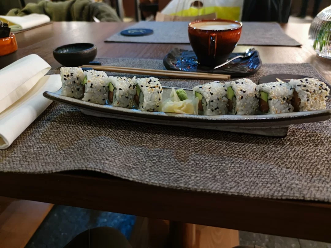 Un plato rectangular con ocho rollos de sushi, jengibre encurtido y wasabi está colocado sobre un mantel individual en la mesa de un restaurante de Mallorca, ofreciendo un toque de lujo. Al fondo, una taza y un plato.