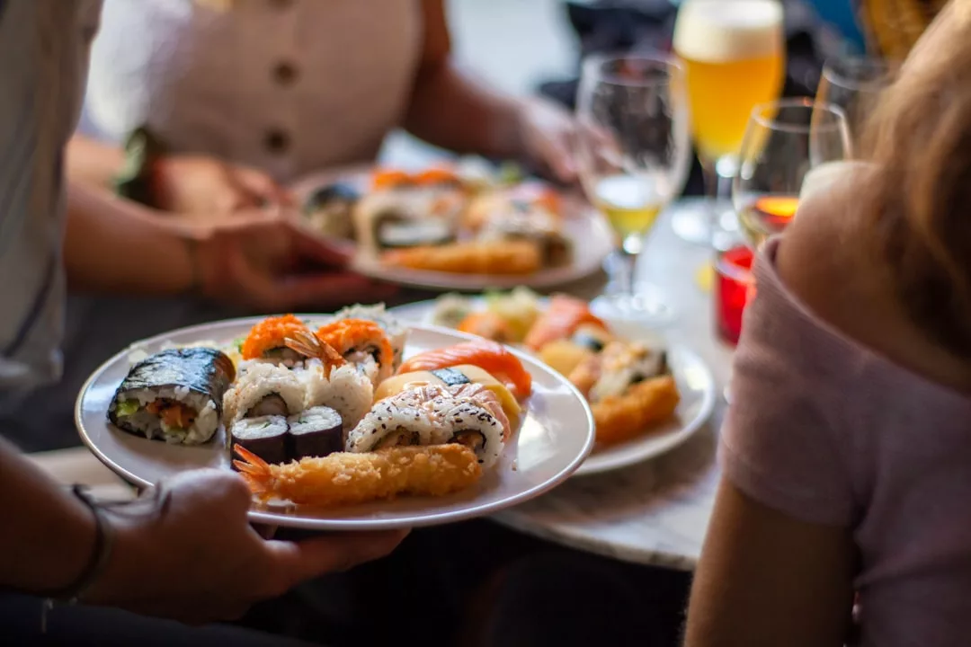 Una persona sirve un plato de sushi variado y tempura en una mesa con bebidas y comida, aportando un toque de Lujo a la experiencia gastronómica en Mallorca, mientras otros están sentados cerca.