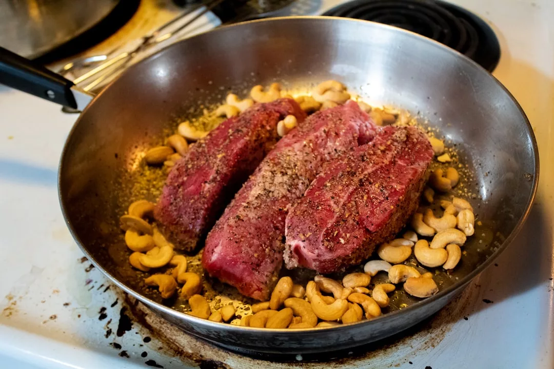 Carne cruda sazonada y anacardos chisporrotean en una sartén de acero inoxidable sobre un hornillo, aportando a su cocina un toque de sabor inspirado en Lujo que recuerda a Mallorca.