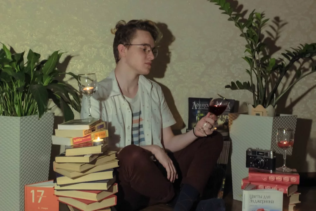 Una persona se sienta en el suelo entre macetas, rodeada de libros apilados y una cámara, saboreando una copa de vino y disfrutando de un momento de lujo inspirado en la relajada elegancia de Mallorca.