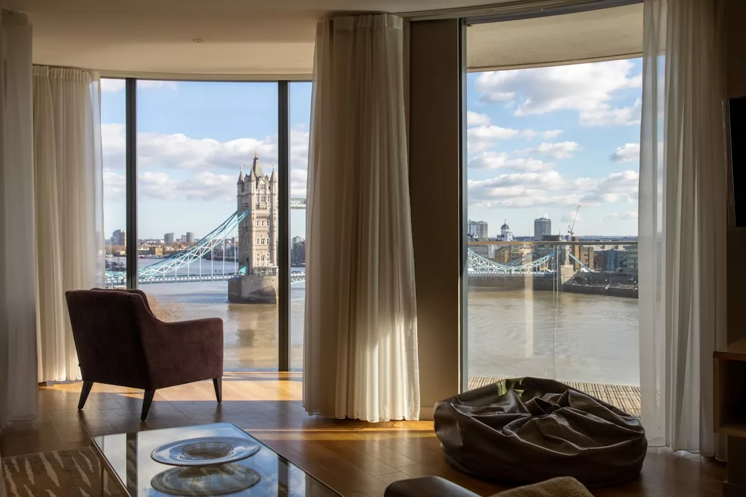 Una moderna habitación con grandes ventanales da al río Támesis y al Tower Bridge de Londres en un día parcialmente nublado. Una silla y un puf ofrecen un toque de lujo, invitando a relajarse y disfrutar de la impresionante vista.