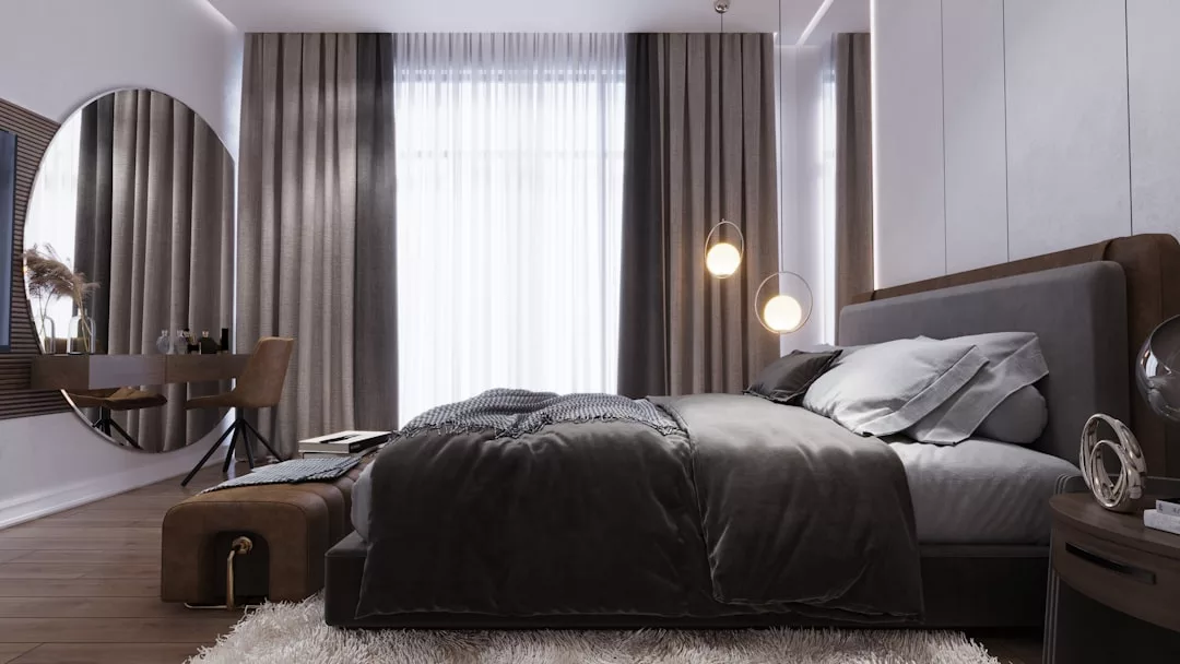 Dormitorio moderno con una cama grande, ropa de cama gris y dos lámparas colgantes. Un escritorio con silla, un espejo redondo y cortinas del suelo al techo completan este refugio de lujo y confort inspirado en Mallorca.