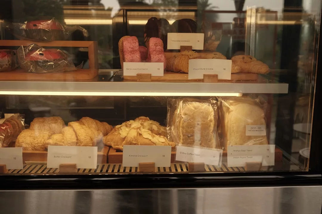 Una vitrina de panadería con diversos productos de pastelería, como cruasanes, pan y galletas, cada uno de ellos etiquetado con su nombre y precio, lo que aporta un toque de Lujo de Mallorca a cada delicioso manjar.